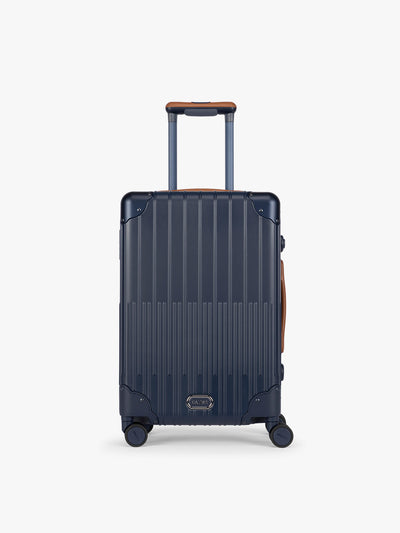 CALPAK Vault Navy blue hard-shell spinner Carry On Luggage with a telescoping handle extended, featuring a brown leatherette side handle and trim, set against a plain light gray background; LVT1020-NAVY view 1