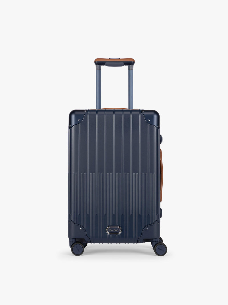 CALPAK Vault Navy blue hard-shell spinner Carry On Luggage with a telescoping handle extended, featuring a brown leatherette side handle and trim, set against a plain light gray background