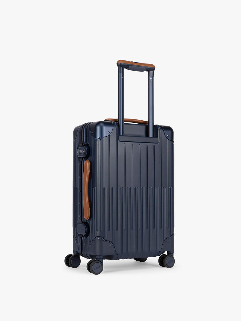 Backside of CALPAK Vault Navy blue hard-shell spinner Carry On Luggage with a telescoping handle extended, featuring a brown leatherette side handle and trim, set against a plain light gray background