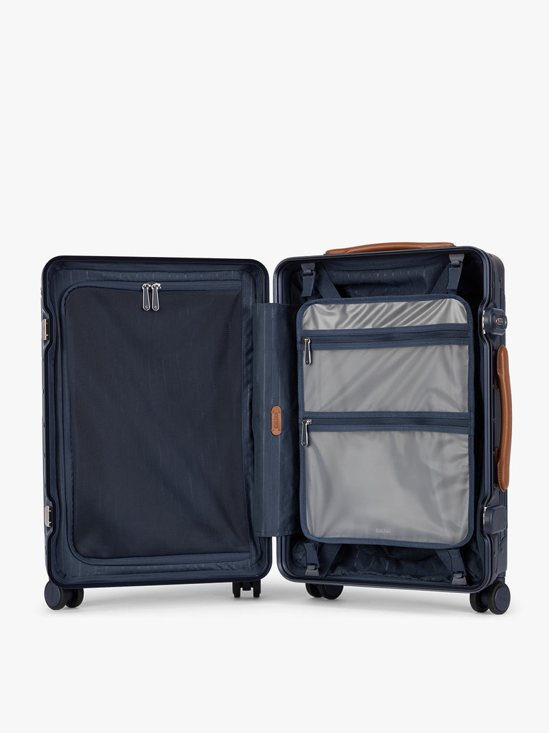 Opened and showing the grey zipper case of CALPAK Vault Navy blue hard-shell spinner Carry On Luggage with a telescoping handle extended, featuring a brown leatherette side handle and trim, set against a plain light gray background