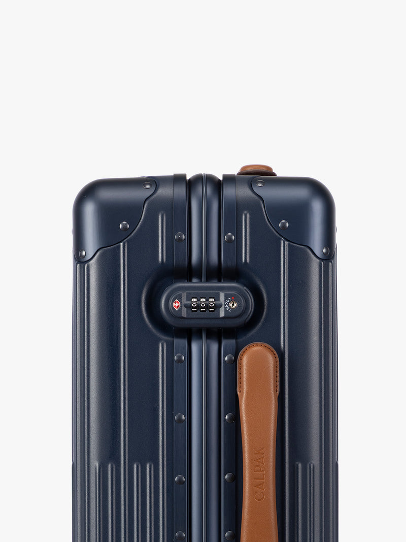 Closeup of side TSA lock of CALPAK Vault Navy blue hard-shell spinner Carry On Luggage with a telescoping handle extended, featuring a brown leatherette side handle and trim, set against a plain light gray background