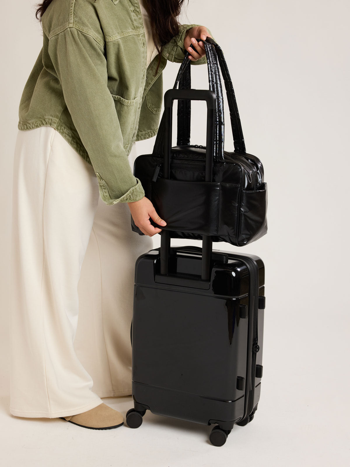 Model slipping on the CALPAK Luka Mini Duffel in shiny Glazed Black through the Trolley Sleeve onto the CALPAK Hue Carry On in Glazed Black; DLM2501-GLAZED-BLACK