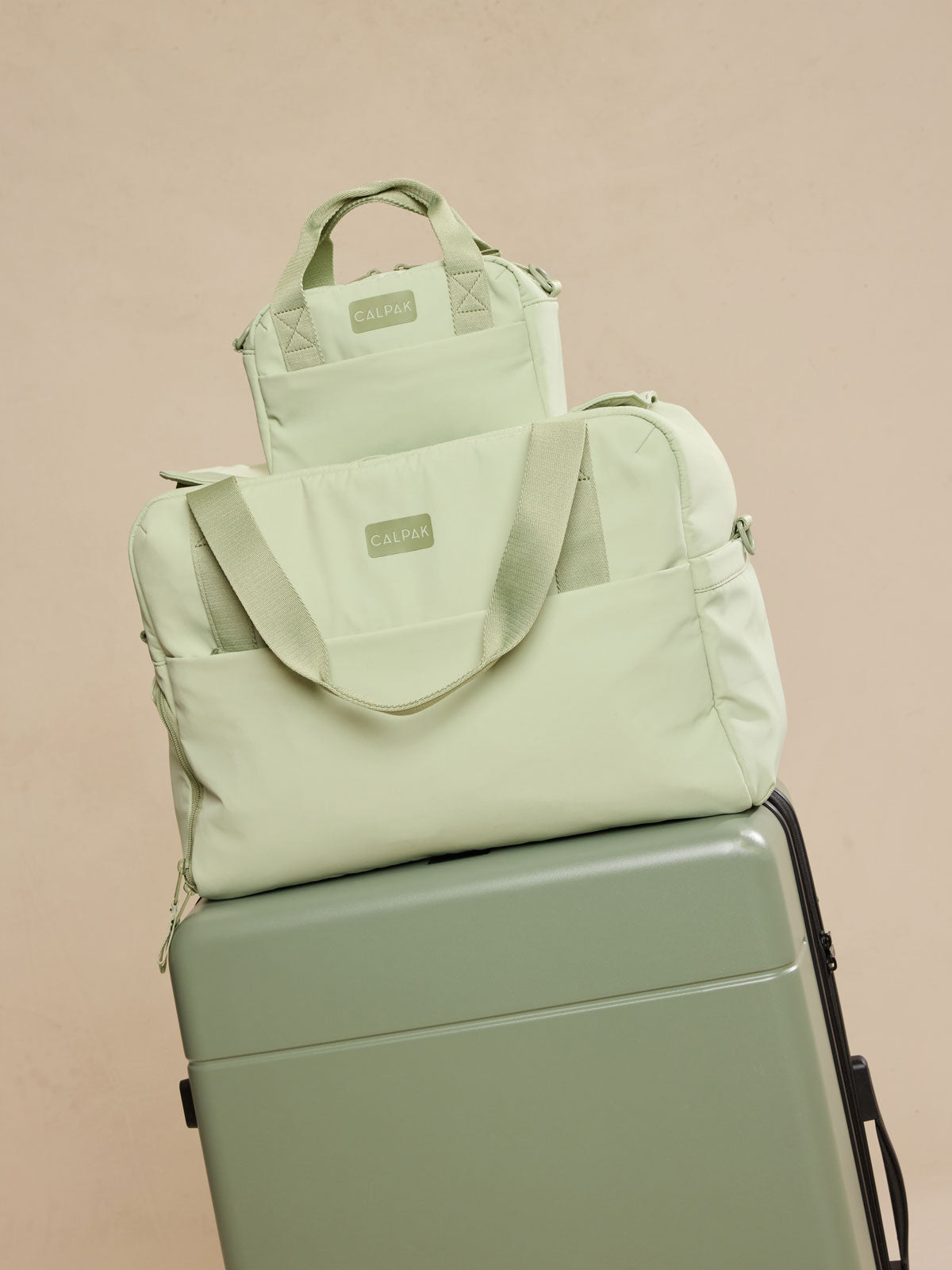 CALPAK Connect Crossbody Bag with adjustable strap in light green Meadow on the CALPAK Laptop Duffel in light green Meadow on a green suitcase on beige background ; CCN2501-MEADOW