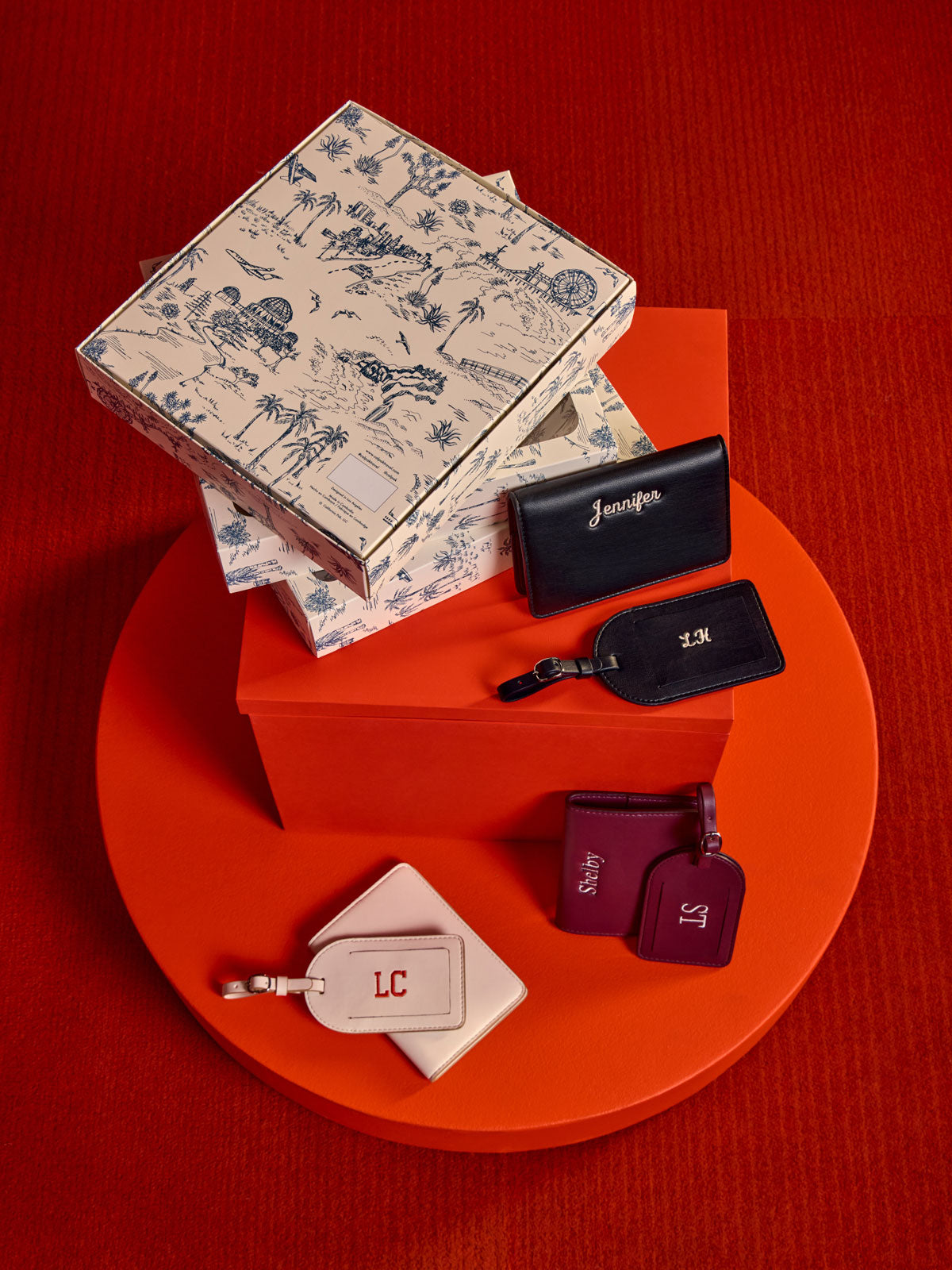 A styled flat-lay of CALPAK Luggage Tag and Passport Holder Set. Showing gift boxes arranged on a circular red platform against a textured red background. The set includes a cream toile-print box, black tags with gold monogram initials, and two burgundy tags with purple monogram initials; PWTS2601-BLACK