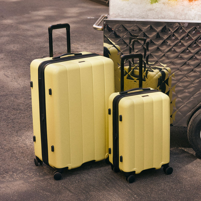 CALPAK Evry Starter Bundle Large and Carry On Luggage in yellow Butter in front of a fruit stand