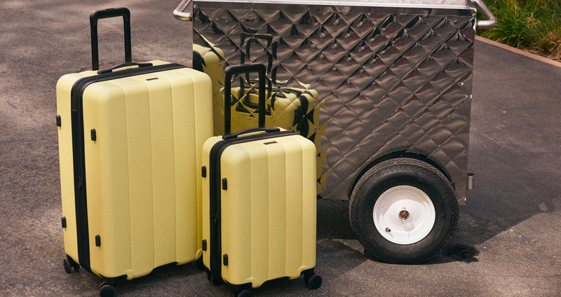CALPAK Evry Starter Bundle Large and Carry On Luggage in yellow Butter in front of a fruit stand