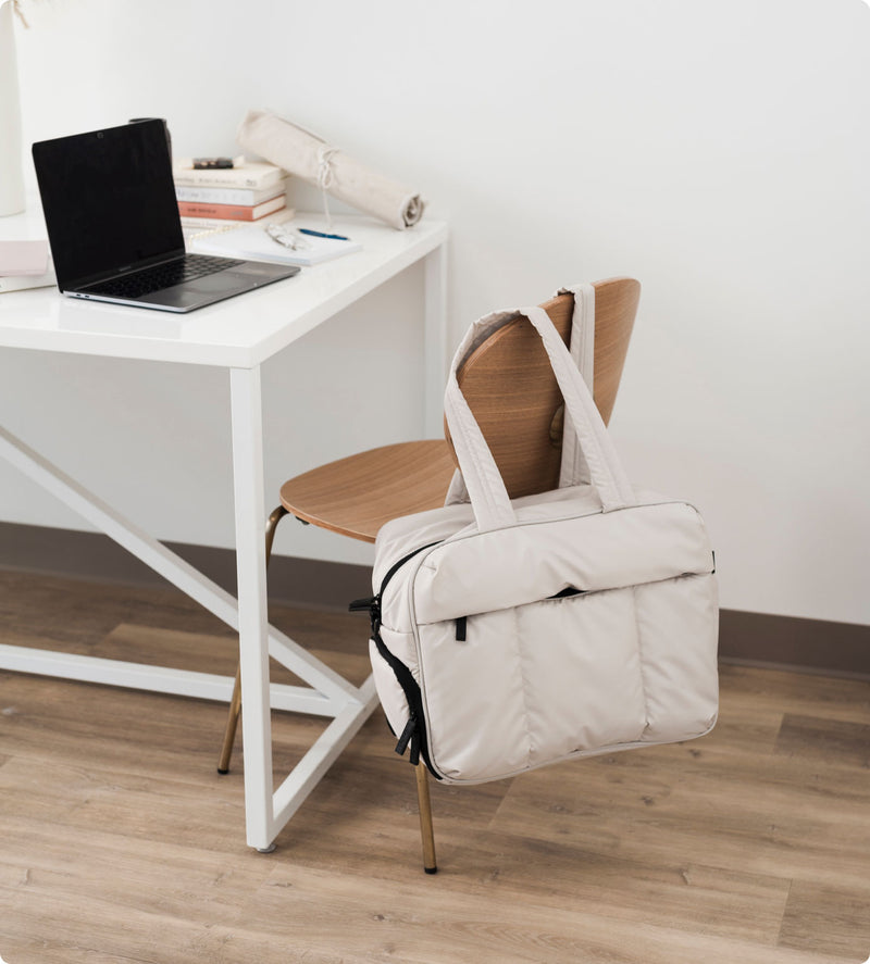 CALPAK ivory Luka Duffel Bag hanging on the back of chair by padded shoulder straps