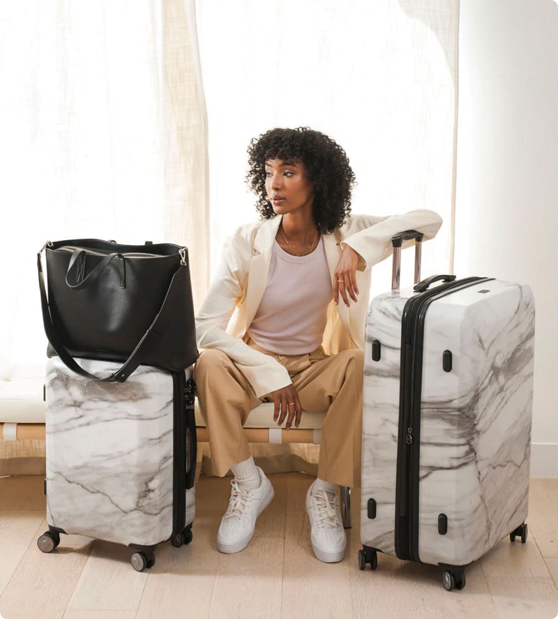 Model sitting with Astyll 2 piece luggage set in white marble and haven tote bag in black