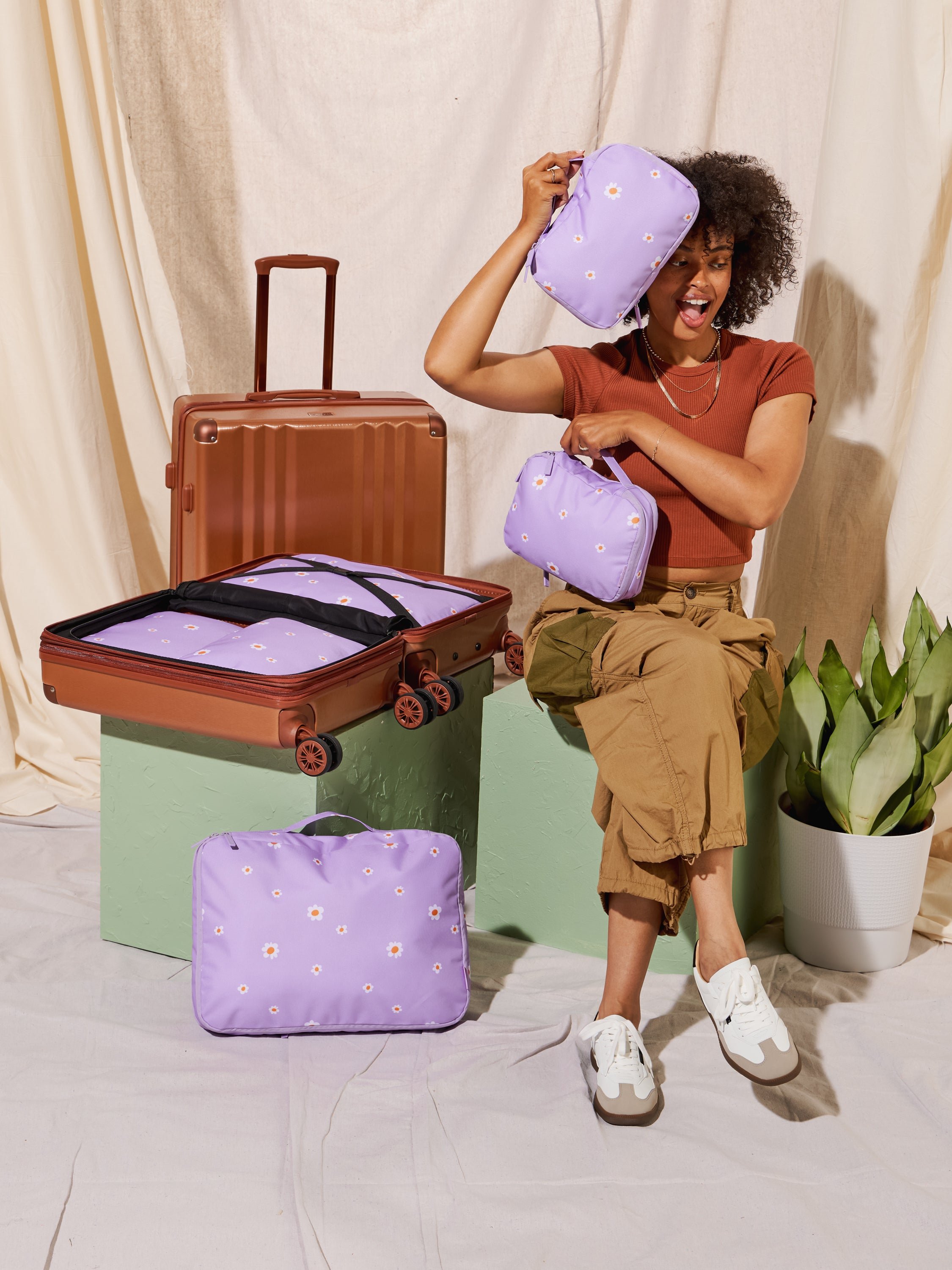 Model holding 2-piece small compression packing cubes in orchid fields among CALPAK Ambeur Carry-On and Medium Luggage in copper, CALPAK Large Compression Packing Cubes and CALPAK Medium Compression Packing Cubes in orchid fields