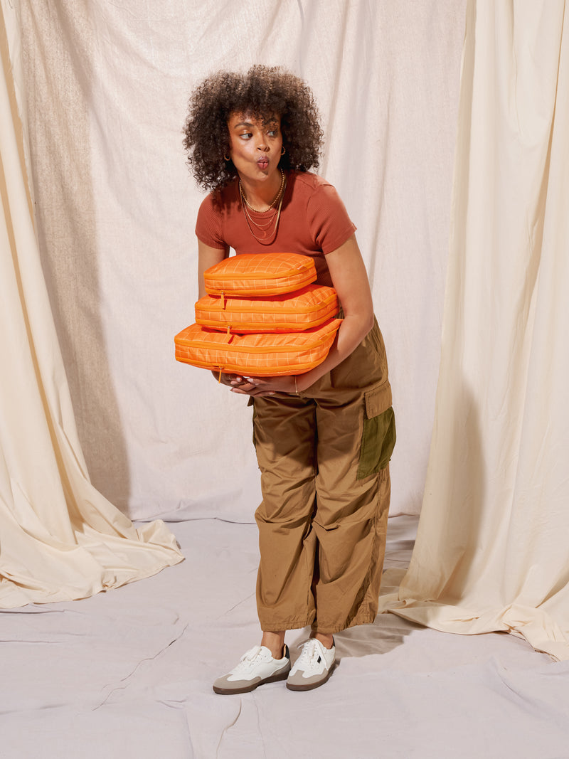 Model holding CALPAK Small Compression Packing Cube, CALPAK Medium Compression Packing Cube, and Large Compression Packing Cube in orange grid for size comparison; PCS2301-ORANGE-GRID