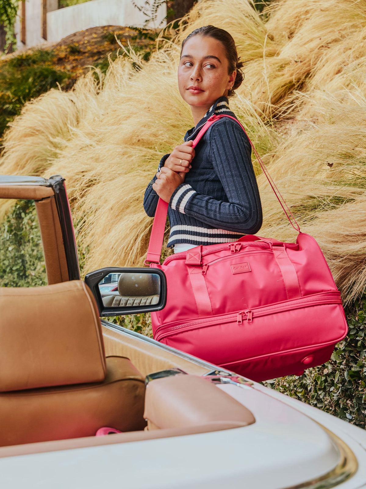 Model standing besides car holding dragonfruit CALPAK Stevyn Duffel with removable strap over shoulder; DST7019-DRAGONFRUIT