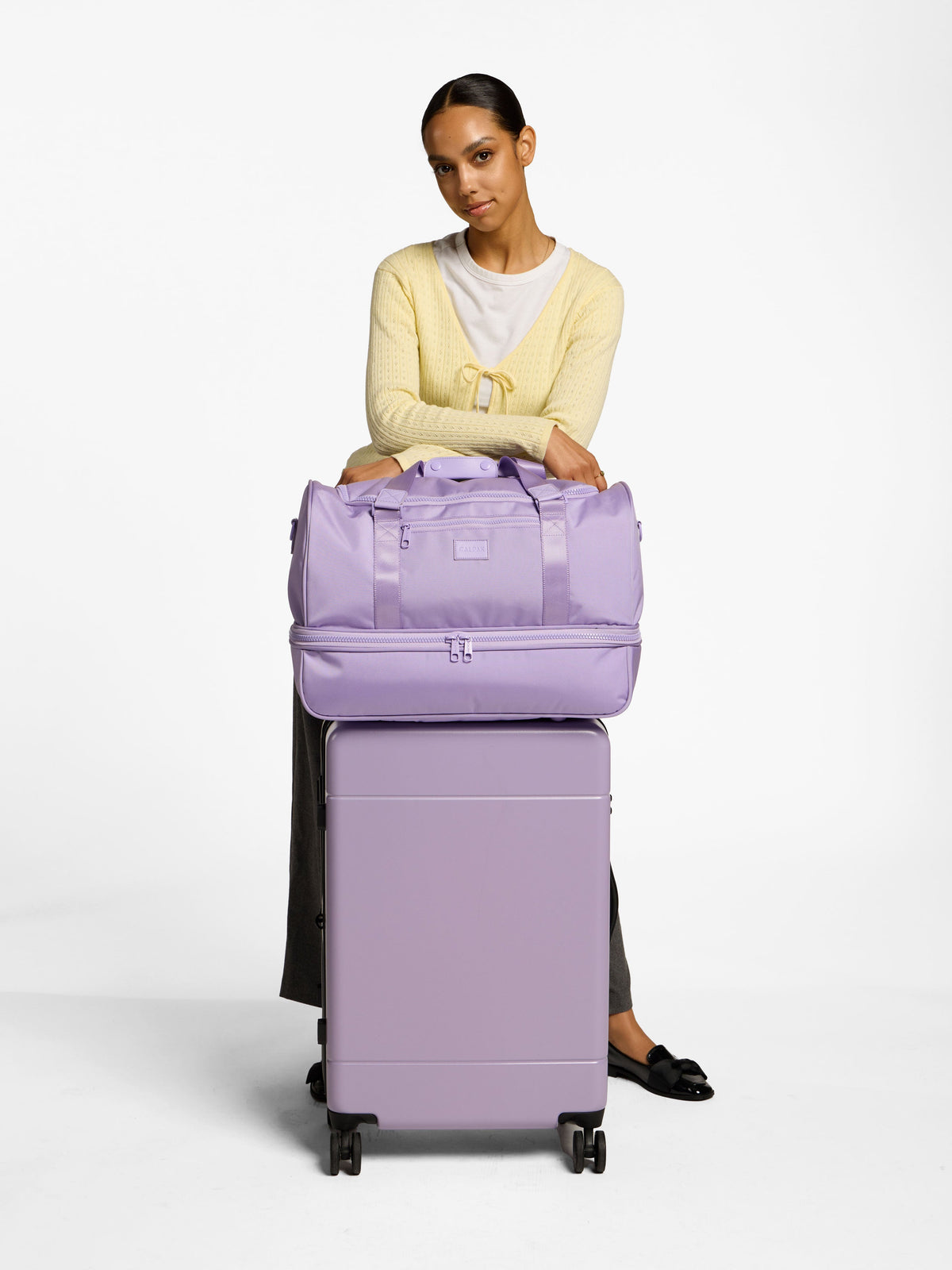 Model standing with CALPAK Stevyn Duffel Bag with Shoe Compartment on top of the Hue Medium Luggage in orchid; DST7019-ORCHID