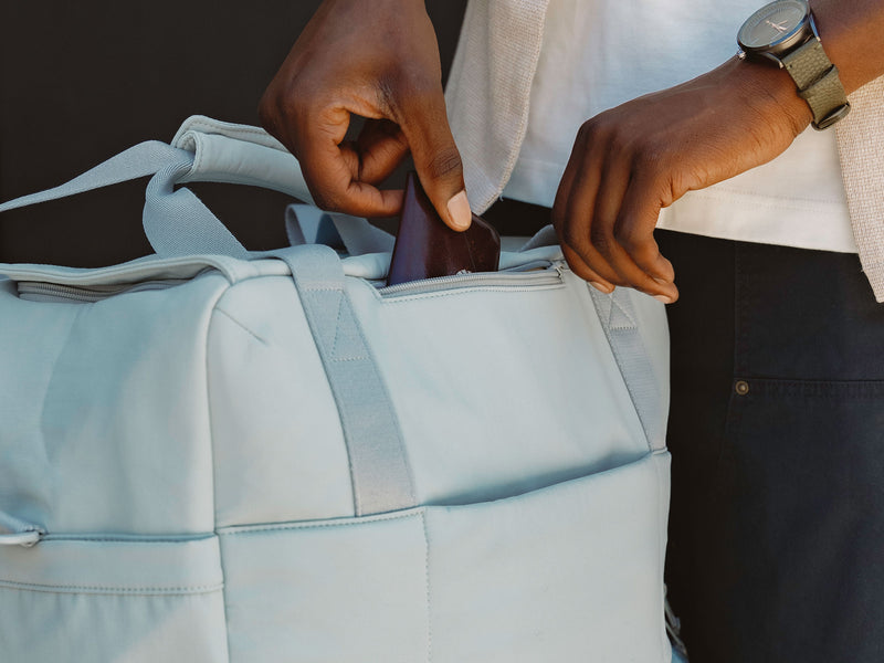 Model placing wallet in front zippered pocket of CALPAK Connect Laptop Duffel in blue Bluebell