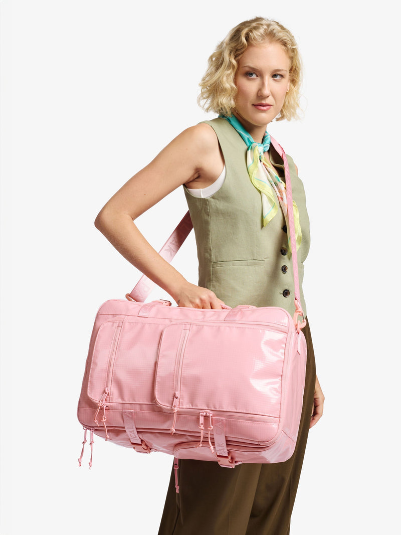 The image features the CALPAK Terra 26L Backpack Duffel in a light pink (blossom) color. It has a soft design with adjustable backpack straps, a top handle, and side handles for versatile carrying.