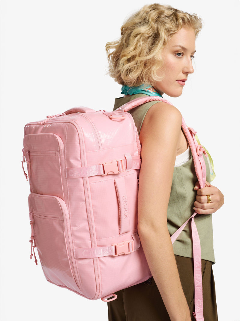 The image features the CALPAK Terra 26L Backpack Duffel in a light pink (blossom) color. It has a soft design with adjustable backpack straps, a top handle, and side handles for versatile carrying.