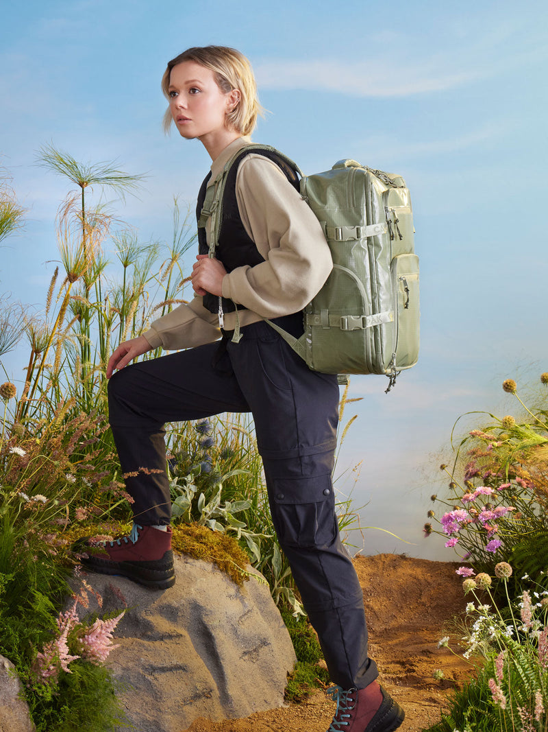 A woman in the foreground wears the Terra 26L Backpack in Juniper is hiking on a rocky outdoor setting background.