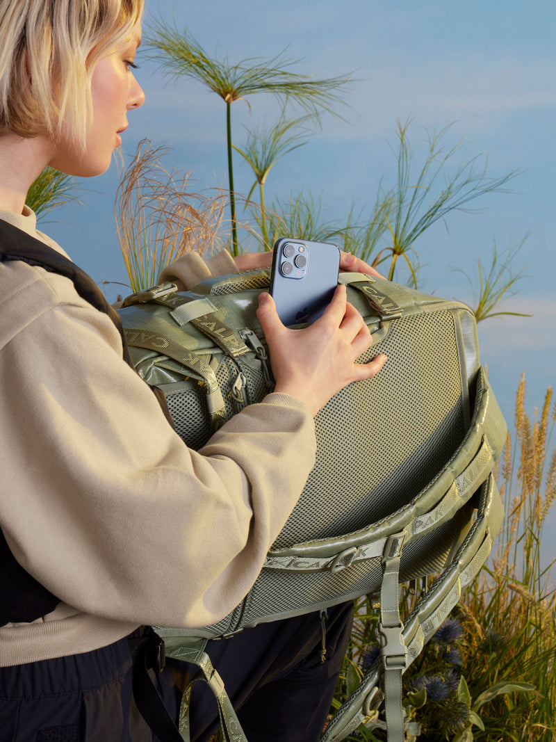  A women in an outdoor setting stands facing away from the camera with the Calpak Terra 26L Backpack. The backpack is shown with a side compartment with the model placing phone inside.