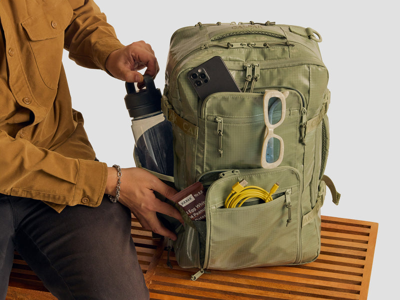 Model packing the Terra 26L Laptop Duffel Backpack in green Juniper with sunglasses, water bottle and other things