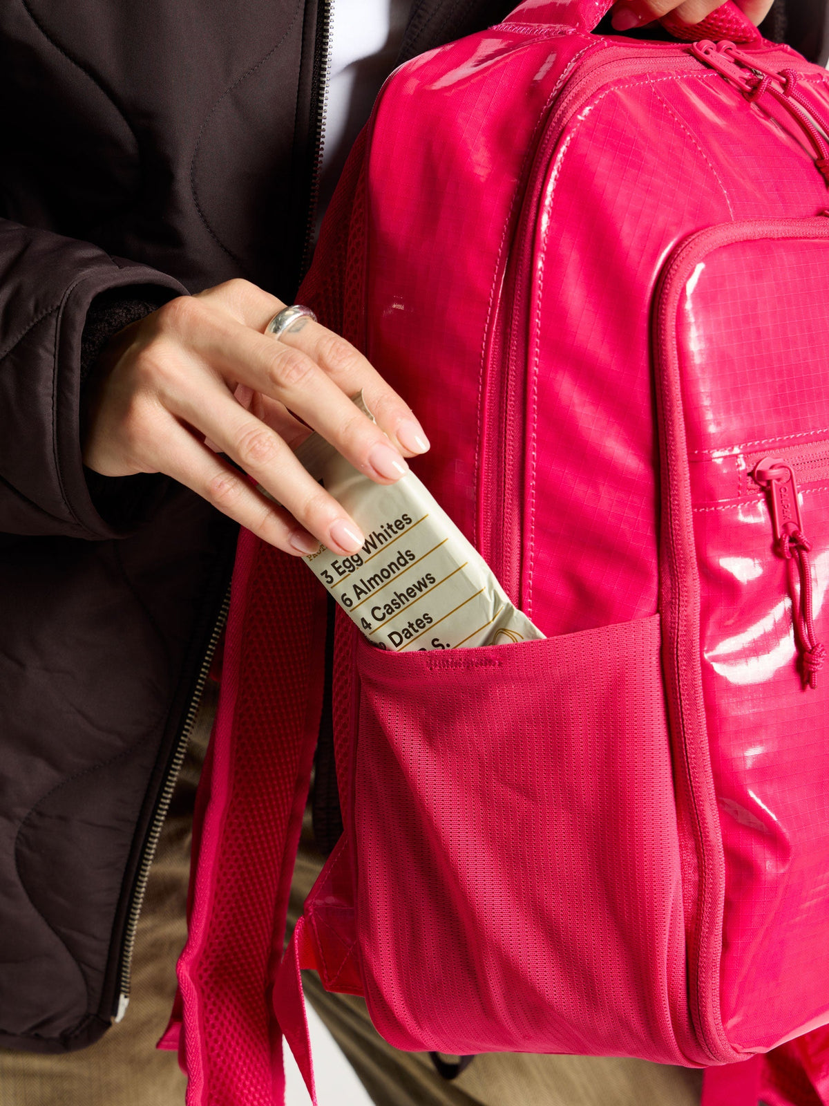 Model placing energy bar within the side pocket of CALPAK Terra Hydration Backpack in dragonfruit; BPT2201-DRAGONFRUIT