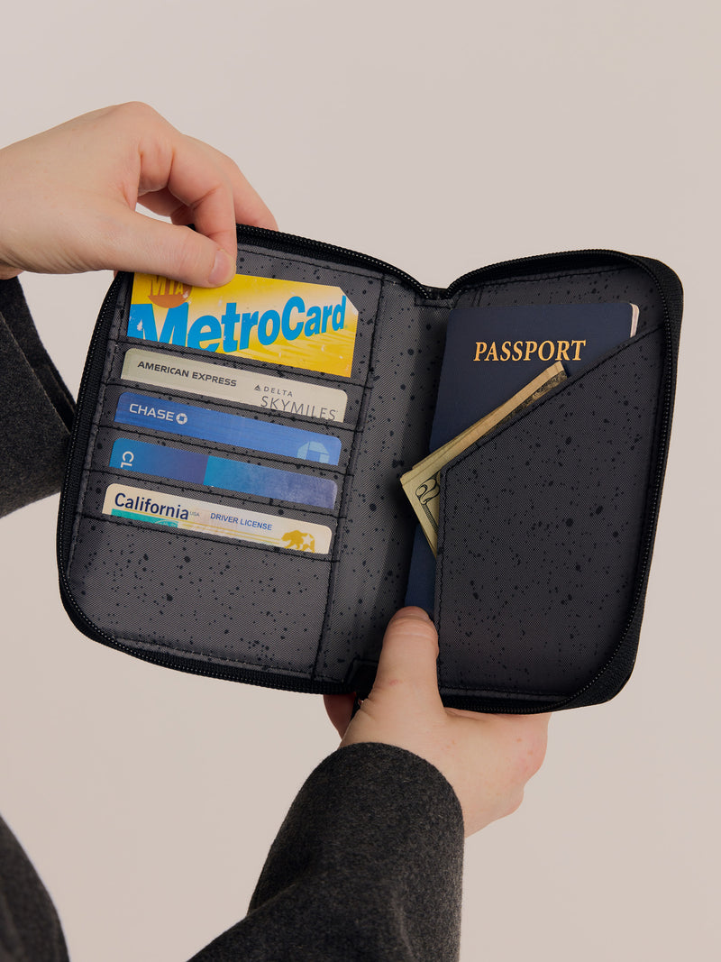 A person holding an open Terra Zippered Passport Wallet in black Obsidian, containing a MetroCard, credit cards, a passport, and cash.