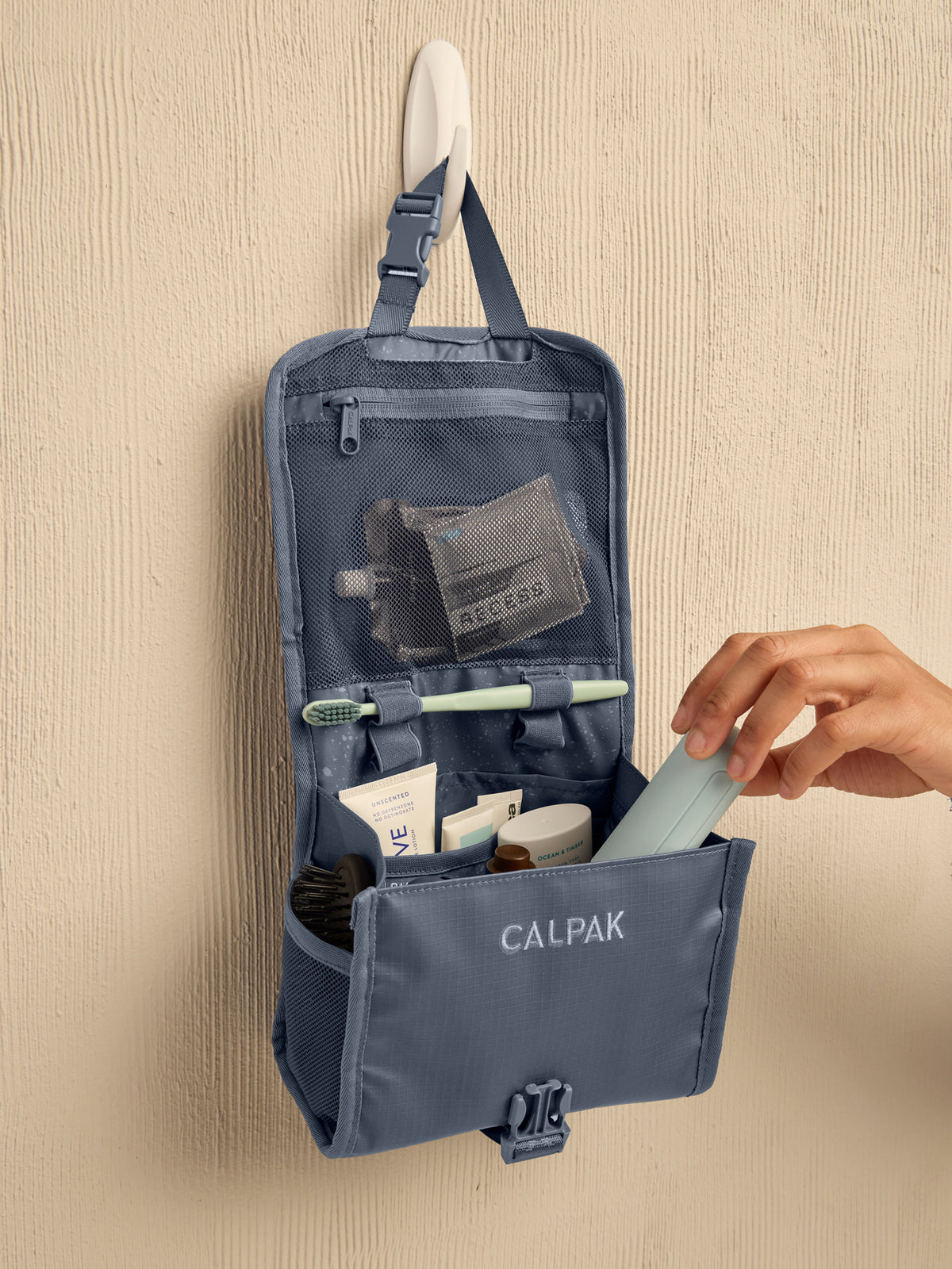Model adding toiletry items to the CALPAK Terra Hanging Toiletry Bag in blue Matte Indigo; THY2201-INDIGO