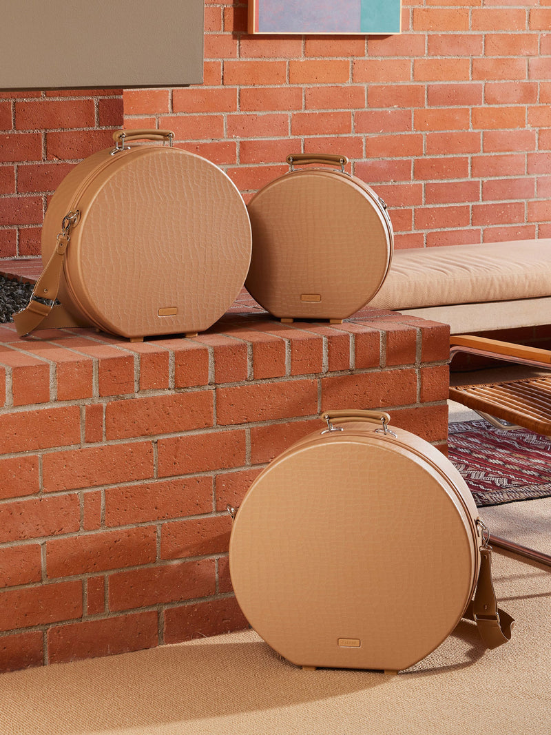 The Trnk Small Hat Box in almond, featuring a structured circular design with a top handle and zippered closure. The case is designed to protect and store hats, making it a fashionable and functional travel accessory