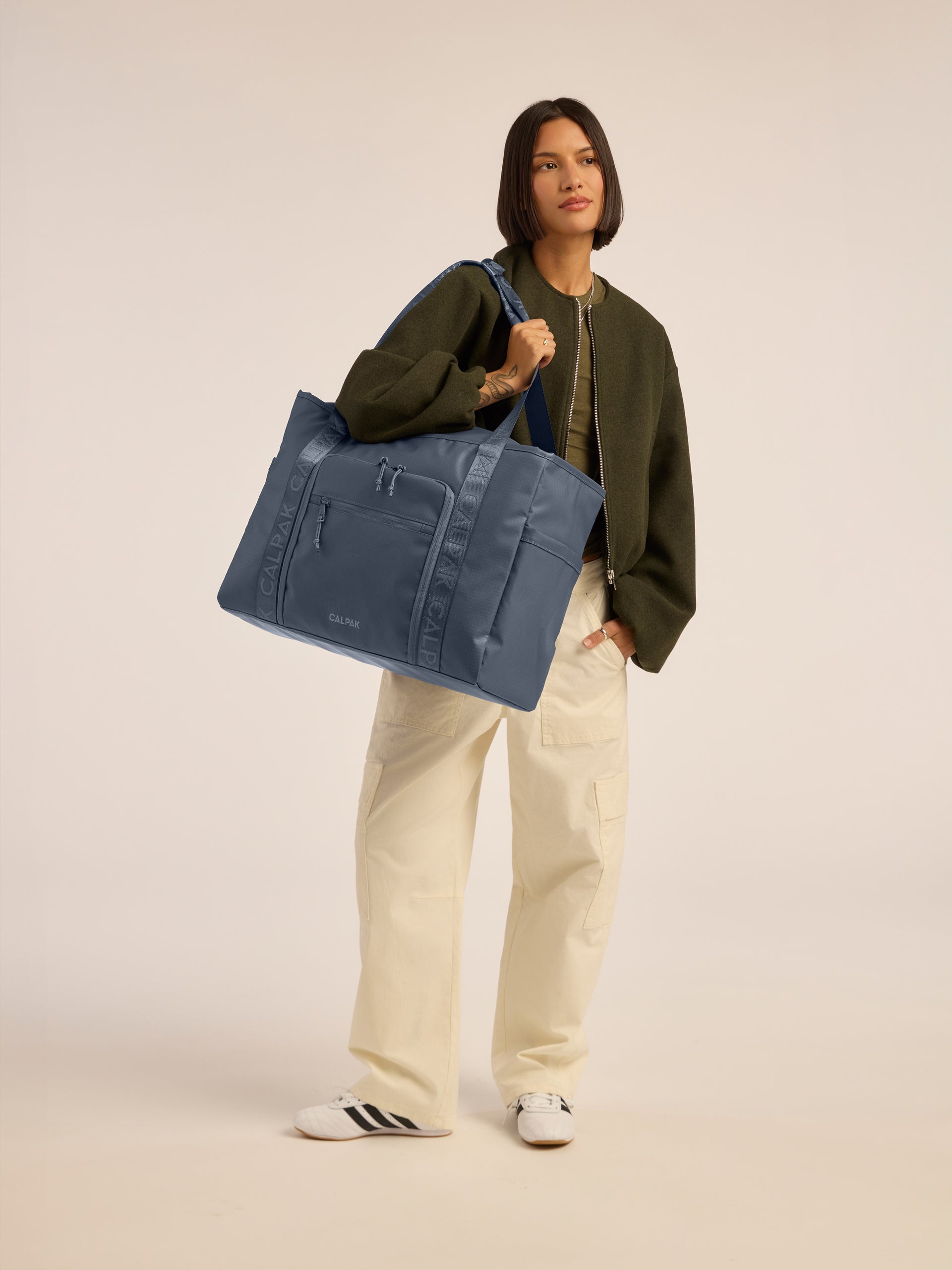 Model wearing CALPAK Terra 35L Water Resistant Zippered Tote Bag in blue Matte Indigo front view; TTO2401-INDIGO
