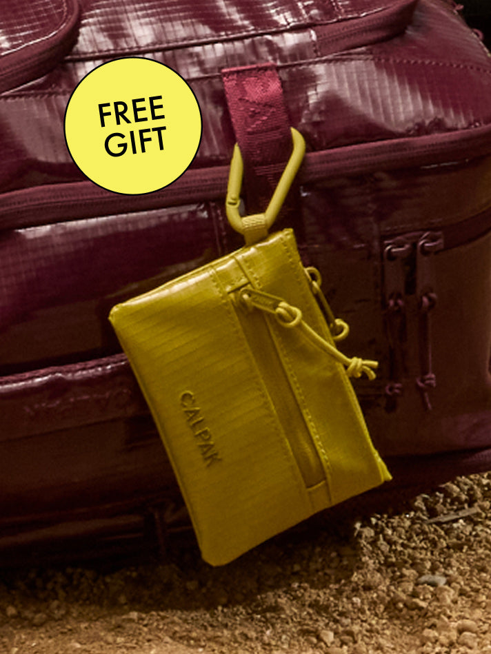 A bright yellow zippered pouch with a drawstring and metal clip, branded CAPPAK, hanging from a dark burgundy textured bag. A yellow circular badge reading 'FREE GIFT' is overlaid in the upper left.