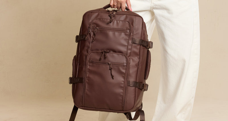 Brown CALPAK Terra 26L Laptop Duffel travel backpack held by a person against a beige background