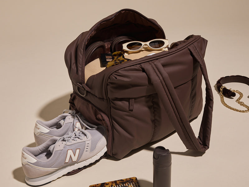 Brown CALPAK Luka Duffel with sunglasses, gray sneakers, and a necklace on a beige background