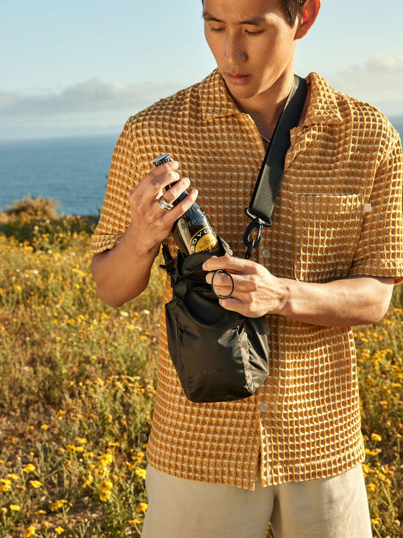 The Water Bottle Holder in black, displayed in a lifestyle setting with a person wearing it over the shoulder with a drink partially inside