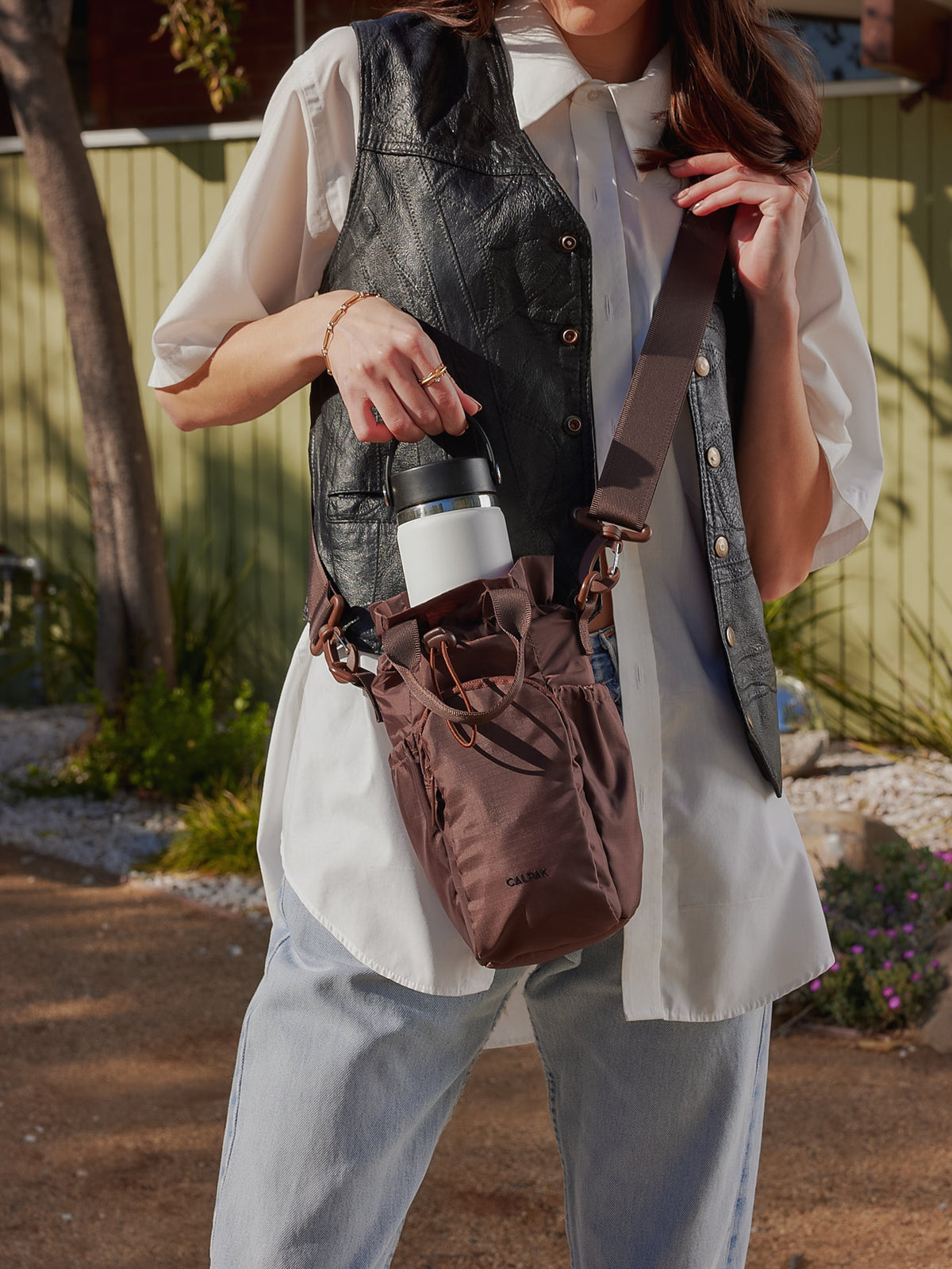Modeling carrying CALPAK Water Bottle carrier with crossbody strap in brown; AWH2101-WALNUT
