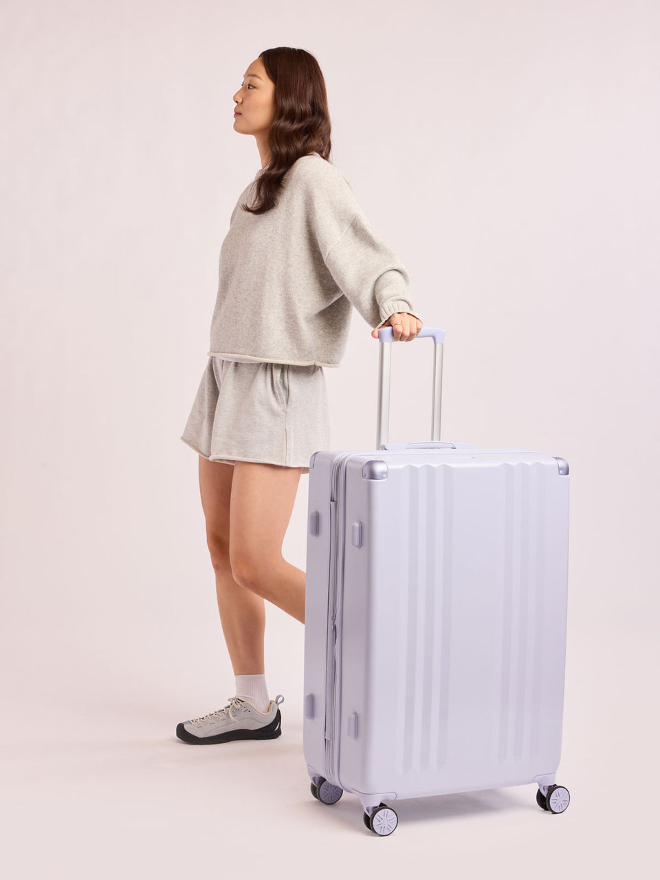 Model in grey with the CALPAK Ambeur Large Luggage in light lavender purple with handles and spinner wheels on a white background; LAM1028-SEA-GLASS