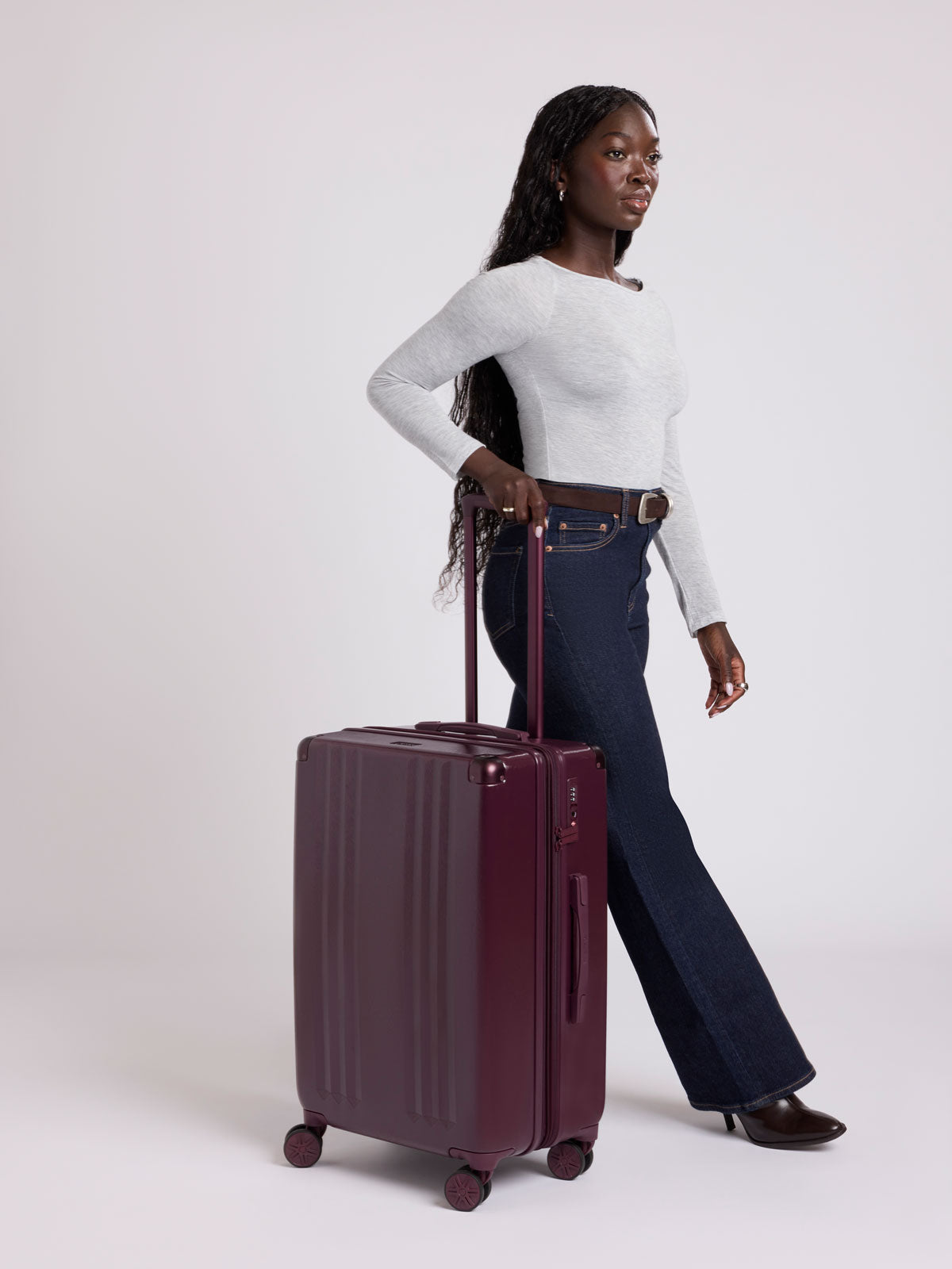Woman in a white shirt and blue jeans rolling the CALPAK Ambeur Medium Luggage in dark burgundy Dark Cherry with vertical ribbed detailing and an extended telescoping handle, shown from the front against a plain white background; LAM1024-DARK-CHERRY