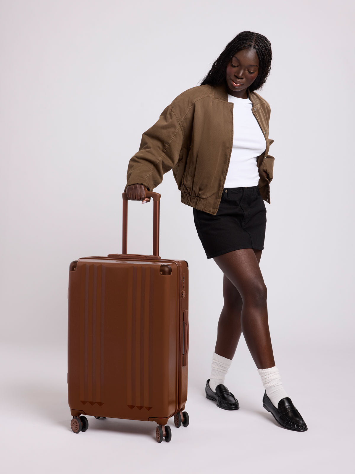 Woman in a white shirt, brown jacket, and black skirt rolling the CALPAK Ambeur Medium Luggage in light Honey Brown with vertical ribbed detailing and an extended telescoping handle, shown from the front against a plain white background; LAM1024-HONEY-BROWN