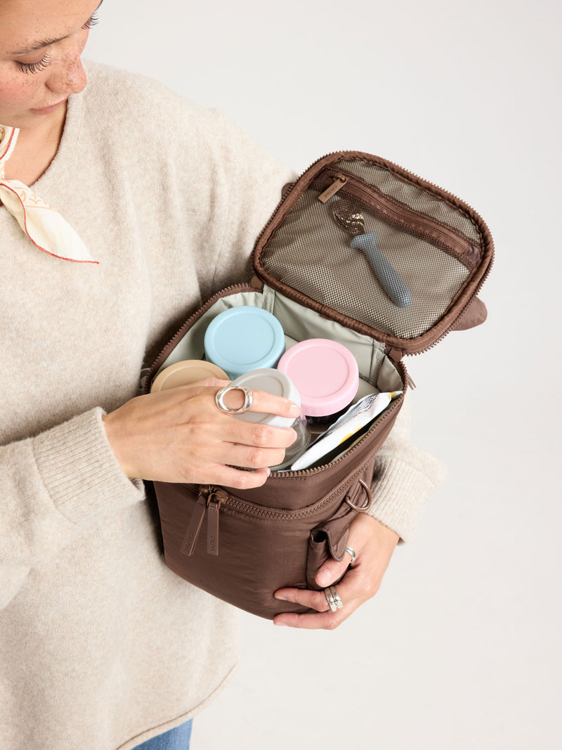 Woman adding items into CALPAK Insulated Baby Bottle Bag in brown Hazelnut