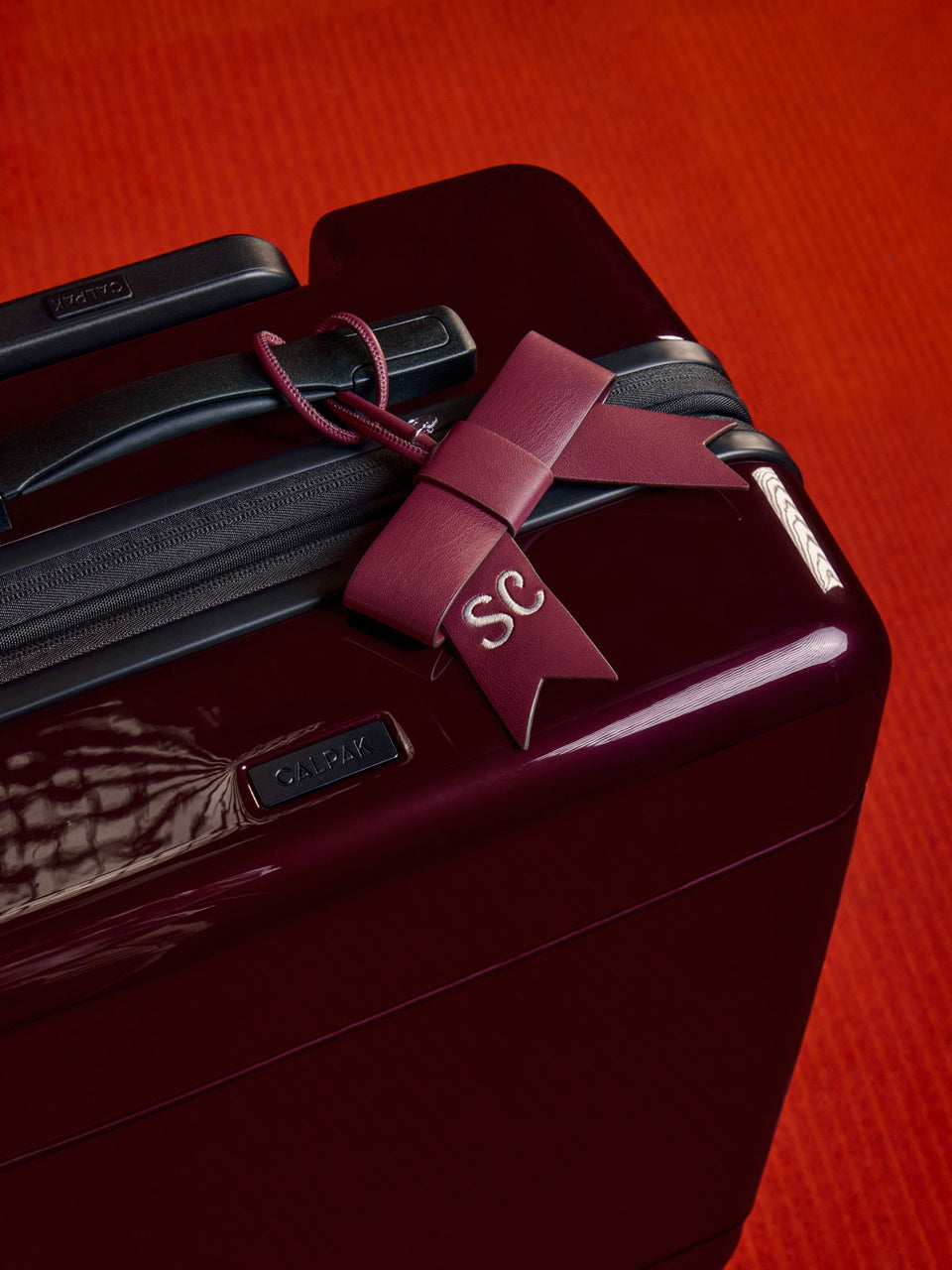 A close-up of a glossy dark burgundy suitcase with a matching burgundy CALPAK Personalized Bow Bag Charm in Dark Cherry tied around the handle, featuring a white embroidered 'SC' monogram and a textured orange-red background; BWBC2601-DARK-CHERRY