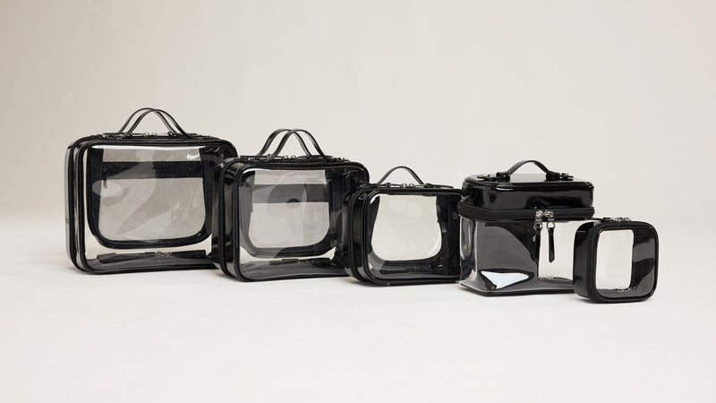CALPAK Clear Cosmetic Cases in sizes Tiny, Small, Medium, and Large with the Clear Train Case as well put side by side for size comparison in color Glazed Black
