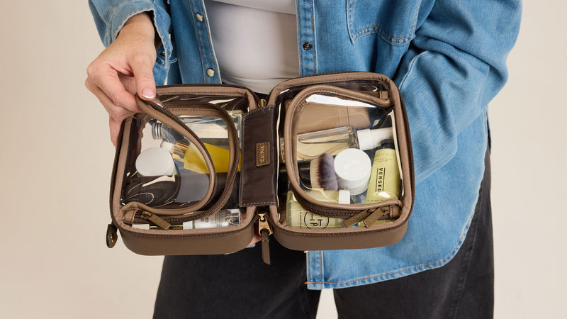 Person holding an open CALPAK Small Clear Cosmetics case  with toiletries inside, wearing a blue denim jacket.