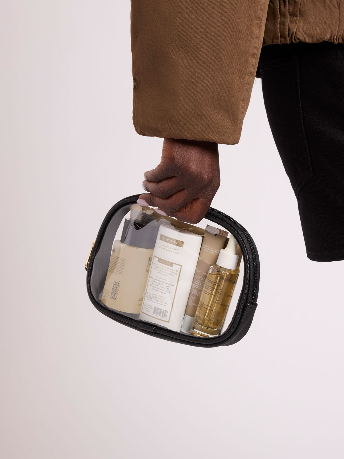 A person in a brown jacket and black pants holds a clear zippered cosmetic pouch with a black trim, containing assorted toiletry bottles and containers, against a plain light background ; CQZP2601-BLACK