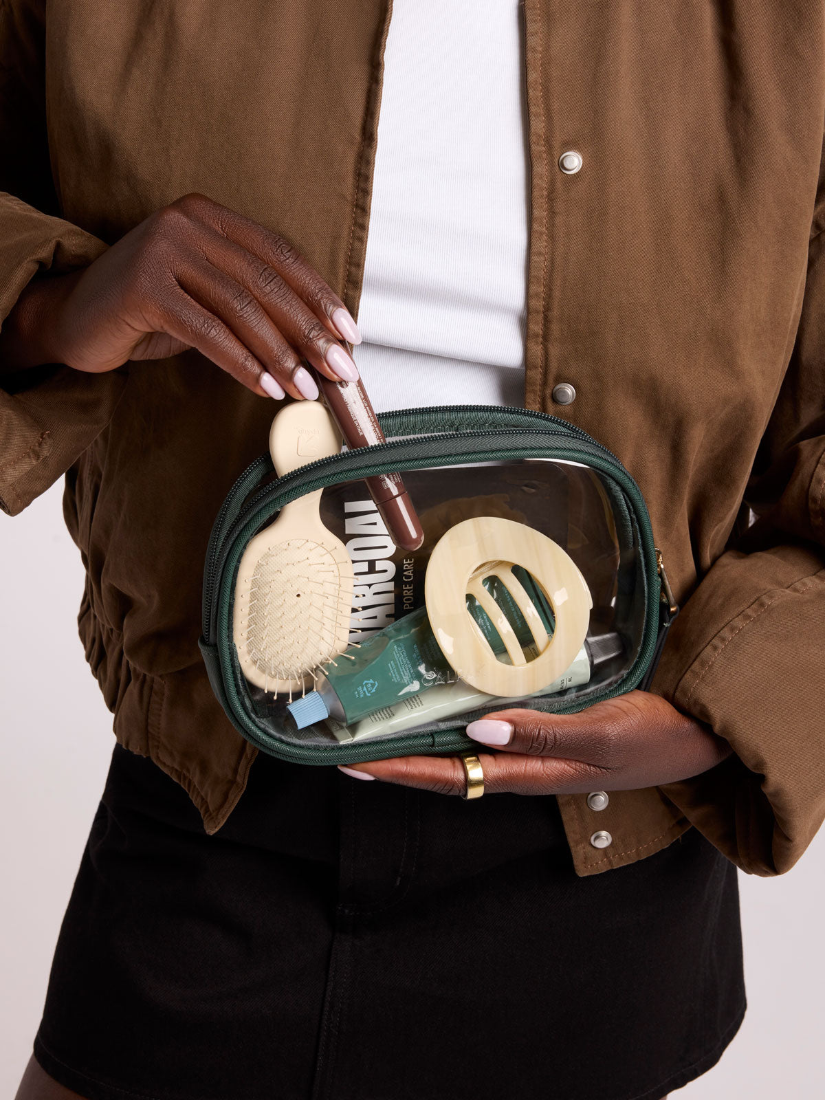 A person holds a dark green transparent zippered cosmetic pouch filled with travel-sized toiletries, including a wooden hairbrush, a small tube, and a round hair tool, against a clean light background; CQZP2601-EMERALD