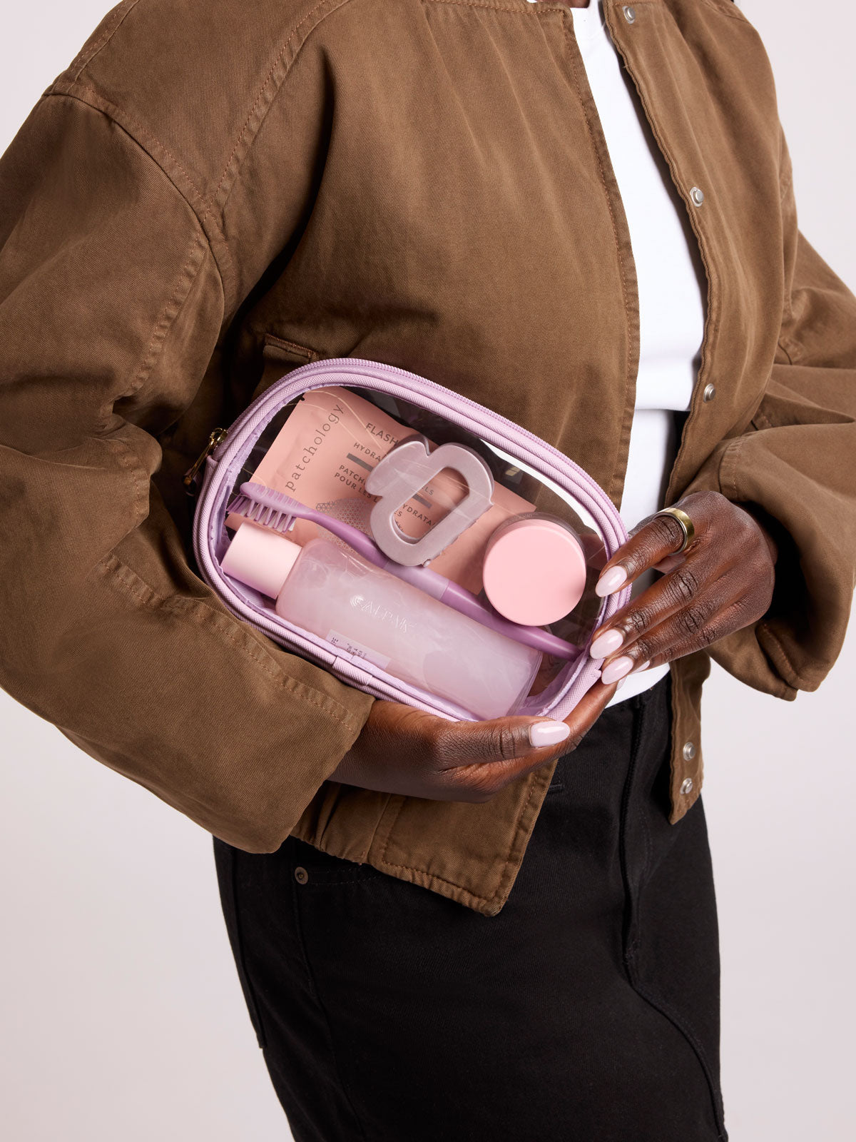 A person wearing a brown jacket and black pants holds a clear zippered cosmetic bag with a lavender zipper ; CQZP2601-LAVENDER