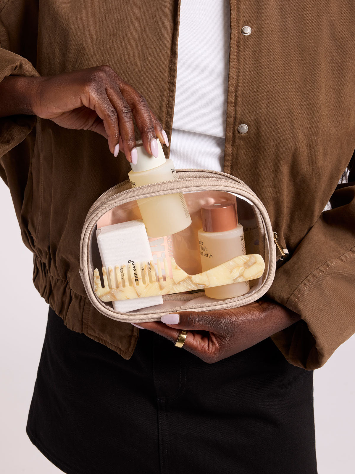A person wearing a brown button-up shirt and black skirt holds a clear zippered cosmetic bag filled with skincare bottles and a cream brush. The person's hands are visible, and the background is a clean white studio backdrop; CQZP2601-STONE