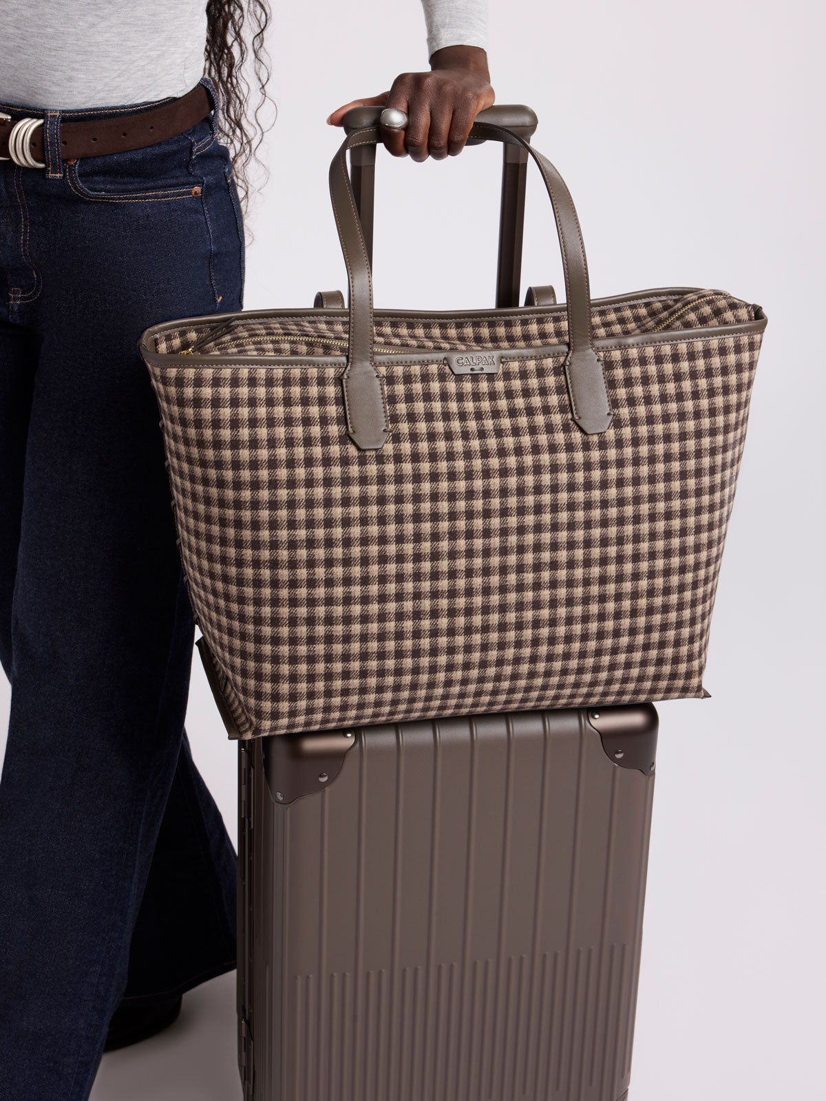 A person wearing dark blue jeans and a light gray top holds a a structured CALPAK Colette 15 Laptop Tote bag with a brown and beige gingham pattern and dark brown leather handles in Mushroom, shown against a plain light gray background; CLCT2601-MUSHROOM-PLAID
