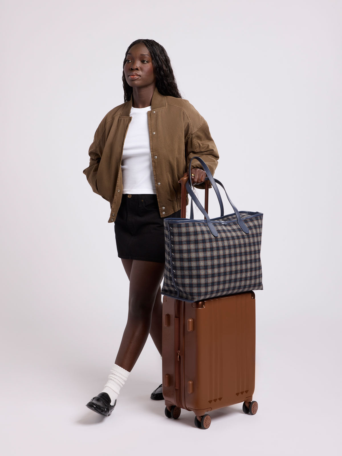 A woman stands in a studio wearing a brown bomber jacket, white top, black skirt, and black loafers, holding a brown rolling suitcase with a CALPAK Colette 15" Laptop tote bag with a brown and navy blue plaid pattern and dark navy blue handles resting on top; CLCT2601-NAVY-PLAID