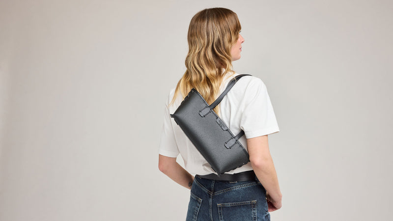 Person wearing a black CALPAK Colette Shoulder Tote handbag with a black strap against a plain background