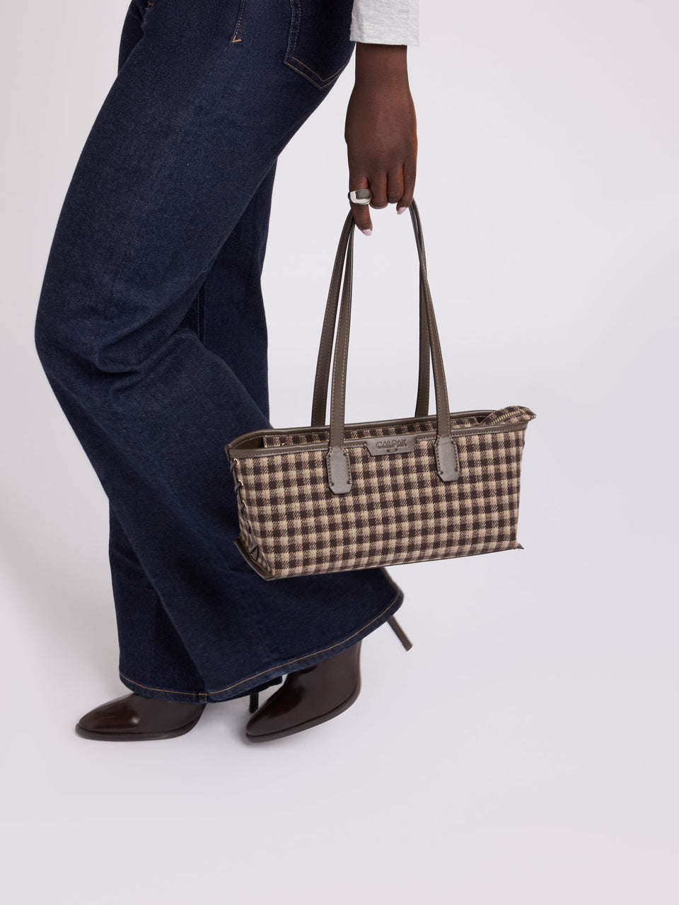 Person wearing dark blue jeans holding the CALPAK Colette Shoulder Tote in Mushroom Plaid featuring a brown and beige gingham check pattern on the body, with dark brown leather handles and trim, shown against a plain light gray background; CLST2601-MUSHROOM-PLAID