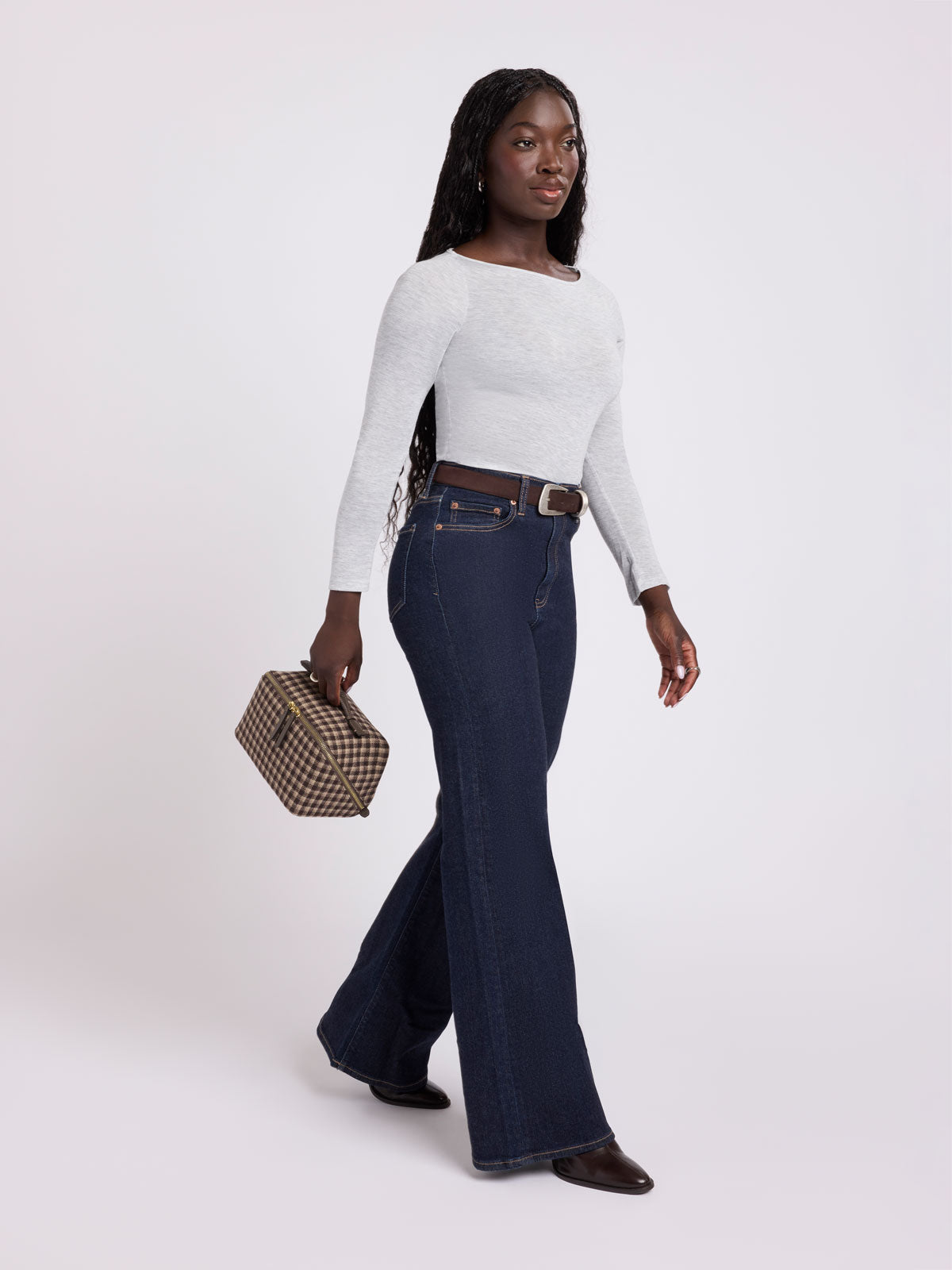 Woman wearing a white shirt and blue jeans carrying the handle of a toiletry bag with zipper and top handle in a grey and brown plaid from CALPAK in Navy Plaid shown against a grey background; CLTB2601-MUSHROOM-PLAID