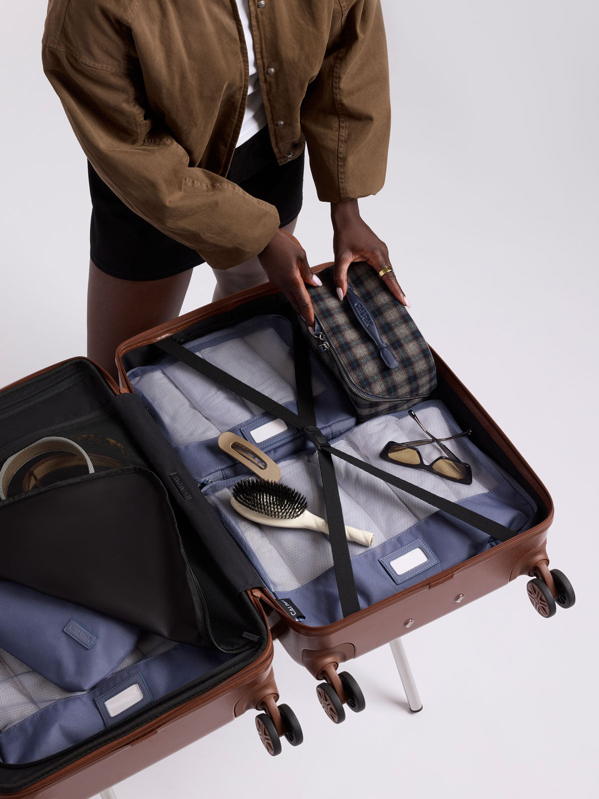 A person packing a toiletry bag with zipper and top handle in a blue plaid from CALPAK in Navy Plaid shown against a grey background; CLTB2601-NAVY-PLAID