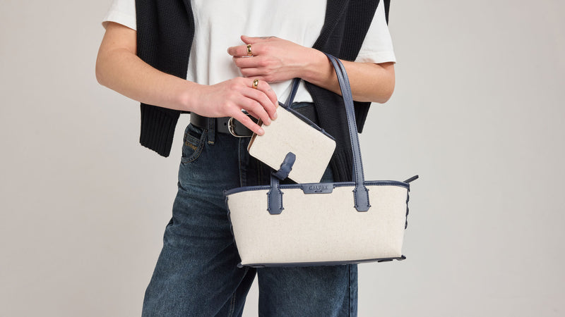 Person holding a cream and blue CALPAK Colette Shoulder Tote in Navy handbag with a gray strap against a plain background